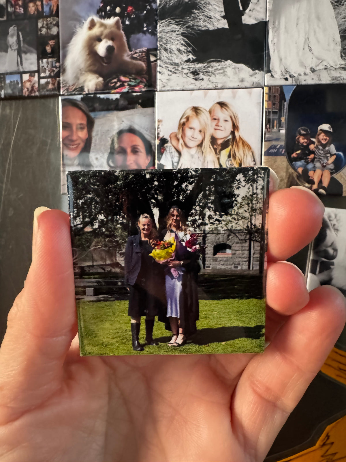 Hand holding a small photo magnet of two people outdoors, with a collage of other photos in the background.